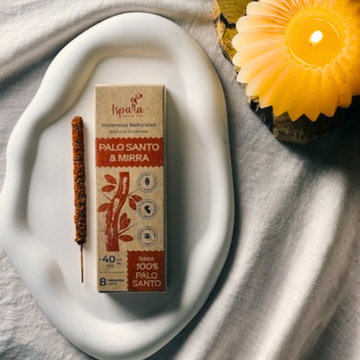Box of Palo Santo and Mirra incense on a white plate with a candle in the background laid on an off white cotton sheet